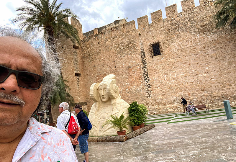 Imagen: Frente a un castillo y de la típica “Dama de Elche”, símbolo de esta ciudad de Alicante, España. Foto de la colección personal del autor.